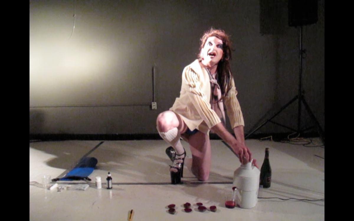 Still from “on the department of experiential medicine II” performance. Crouched on the floor surrounded by a bottle of champagne and enema equipment, Mikiki juices halved beets into a clear cup.