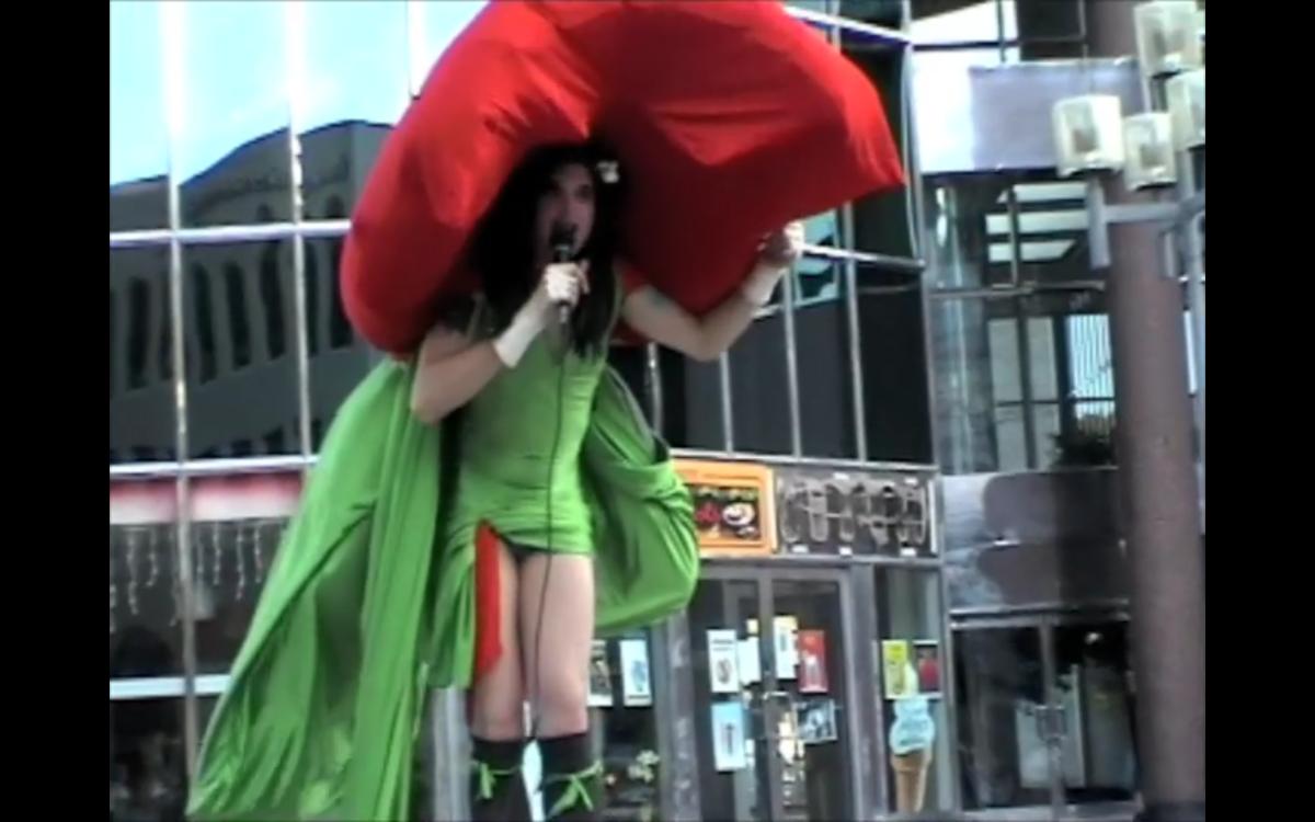 Still from Drag Race X. In a tight green dress with a train, Mikiki’s bright red cape is inflated to flip up over their head as “authoritative shoulder pads” as they speak into a microphone.