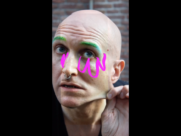 Mikiki, with a shaved head and green eyebrows, pinches and pulls the skin at their jawline, lips slightly parted. Pink swoops are drawn on the photo under their brilliant green-blue eyes.