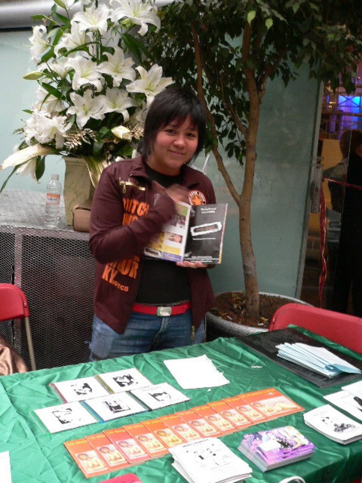 The photo shows a person, Chichis Glam, tending a table full of flyers and other lesbian ephemera at the 2nd Lesbian Festival organized by Martha Cuevas at the Cabaré-Tito VIP in Mexico City. Chichis is holding up a piece of ephemera and looking at the camera.