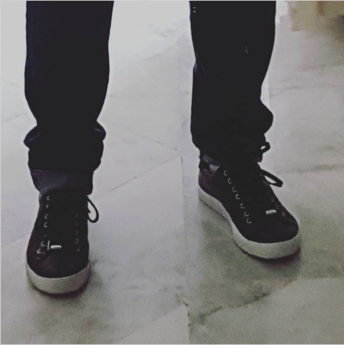 black sneakers, white soles, bottom of legs in black jeans with rolled cuffs, on a grey concrete floor