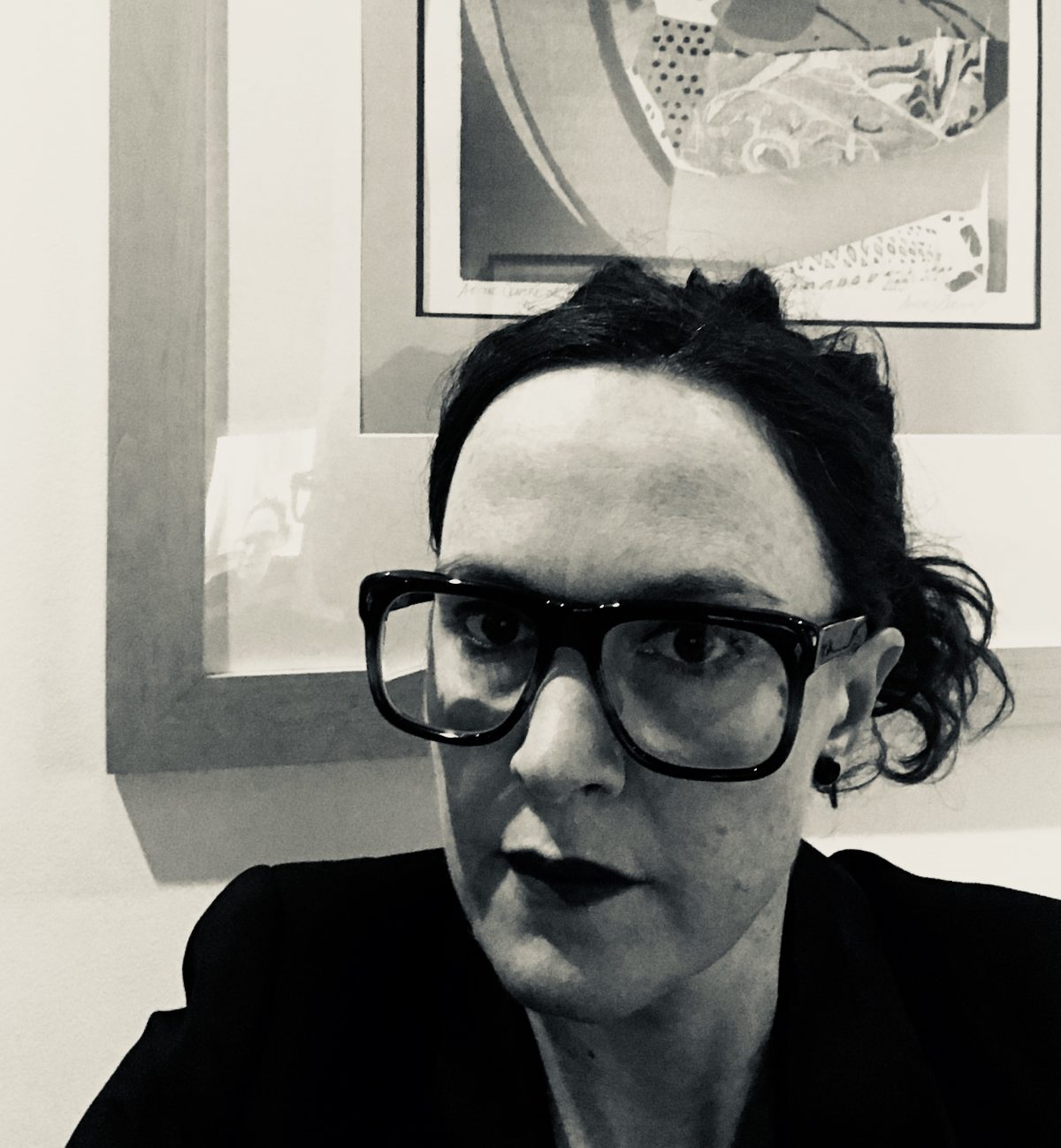 Black & white image of T.L. Cowan. Image shows a white femme wearing dark, thick-rimmed eyeglasses, brown curly hair pulled back into a lose bun; she sports dramatic dark red lipstick. She is wearing a black blazer. The background of the image is the bottom corner of a framed print by Andréa Zarwiny. T.L.'s reflection is mirrored in the glass of the picture.
