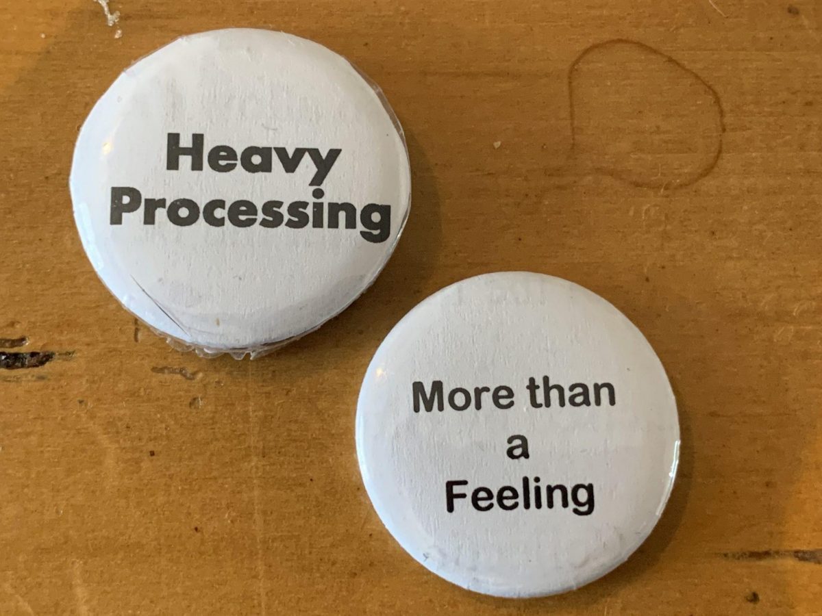 Two home-made buttons — white with black letters. One reads Heavy Processing; the other reads More Than a Feeling. Background is a yellow wooden coffee table with a coffee stain.